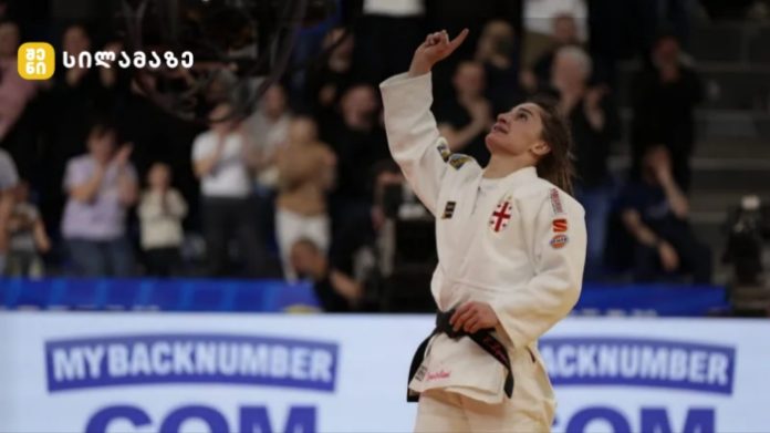 Female judo athlete in a white gi with a black belt raises her right fist in celebration on the mat, with cheering spectators in the background.