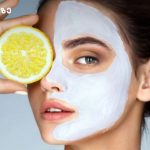 სახის ნიღაბი ციტრუსისგან Woman with a white facial mask holding a lemon slice over her eye during a skincare routine.