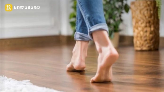 Person walking barefoot on a wooden indoor floor, jeans cuffs rolled up at the ankle.